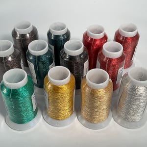 May include: A collection of twelve metallic embroidery thread spools arranged in three rows. The spools feature vibrant colors including red, green, gold, silver, dark brown, and teal, each wound on a white plastic core with a white top cap.