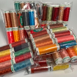 May include: A collection of assorted sewing thread spools in various colors including red, orange, yellow, green, blue, and pink. The spools are packaged in clear plastic bags, with some labels visible showing text such as Maschinengarn and Stopfgarn.