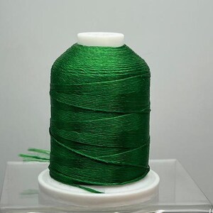 May include: A spool of vibrant green thread, tightly wound around a white plastic base. The thread has a slight sheen, and a few loose strands are visible. Ideal for sewing, embroidery, or crafting projects.