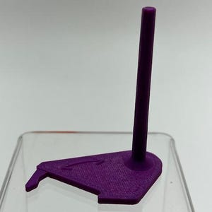 May include: A purple 3D printed display stand featuring a flat, triangular base with a small protruding hook on one side and a single, vertical cylindrical post extending upward from the center of the base for holding items in an upright position.
