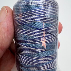 May include: A spool of variegated thread wrapped in clear plastic film, featuring a multi-colored pattern of blue, light blue, and silver-grey strands wound in a crisscross design with a small orange mark on the surface.