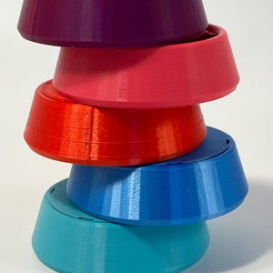 May include: A stack of five colorful, conical objects in shades of purple, pink, red, blue, and teal. Each object has a flat top and a flared, ridged base. The objects are arranged in a vertical tower, with each one slightly offset.