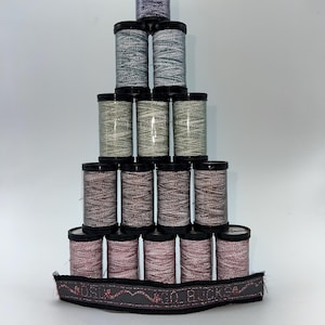 May include: A pyramid of spools of thread in various colors, including purple, gray, and pink, with black lids. A dark gray ribbon with red and pink accents and the text "OKO BUCKS" is at the base.