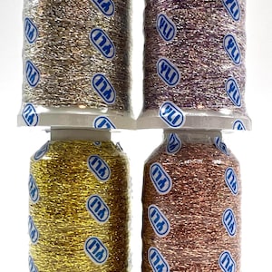 May include: Four spools of metallic thread in gold, bronze, silver, and purple. Each spool is wrapped in clear plastic with the "YLI" logo. The thread is tightly wound, and the colors shimmer.