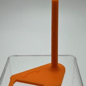 May include: An orange plastic stand with a vertical post and a triangular base. The base has an arrow symbol and a small lip on one side. The stand is designed to hold an item upright.