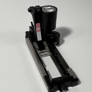May include: A black sewing machine presser foot with a silver metal base. The presser foot has a clear plastic section and a black cylindrical component. The text "3C" is printed on a red label.