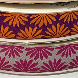 May include: Spools of decorative ribbon in a stack. The top spool features orange floral designs on a purple background. The bottom spool has a pink floral pattern on a gray background. The ribbon is likely used for crafts, gift wrapping, or sewing projects.