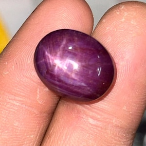 Puede incluir: Una gema pulida, de forma ovalada, de color rojo intenso, con un efecto de estrella. La gema se sostiene en una mano, mostrando su superficie lisa y los juegos de luz internos. El color de la piedra varía de un rojo oscuro y rico a un tono más claro y vibrante.