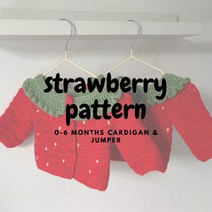 May include: Two red crocheted baby cardigans with green collars, resembling strawberries, hang on a white wall. The cardigans have white dots and are labelled "strawberry pattern" and "0-6 MONTHS CARDIGAN & JUMPER".