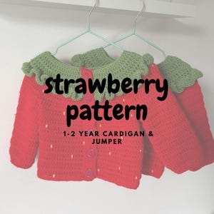 May include: Three red crocheted strawberry-themed cardigans with green collars and button fastenings hang on a rack. The text "strawberry pattern" and "1-2 YEAR CARDIGAN & JUMPER" are displayed on the front of the cardigans.