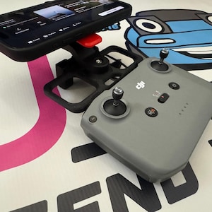 Pode incluir: Um smartphone preto está montado em um suporte de controlador de drone preto e vermelho. Um controlador de drone cinza com joysticks e botões está abaixo do suporte do telefone. O fundo apresenta um gráfico de desenho animado de um carro.