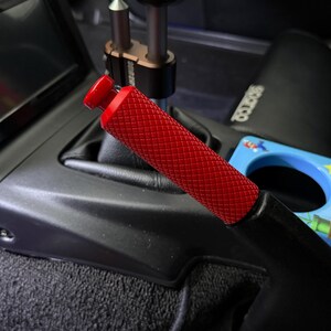Könnte beinhalten: Ein roter Handbremsgriff aus Metall mit einer texturierten, rautenförmig gerändelten Grifffläche. Der Griff ist im Innenraum eines Fahrzeugs neben einer schwarzen Mittelkonsole, einer Gangschaltung und einem blauen Getränkehalter montiert.