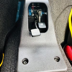 Könnte beinhalten: Eine schwarze Kunststoffplatte mit zwei Schrauben und einer rechteckigen Öffnung, die einen Metallhebel und einen schwarzen Knopf freigibt.