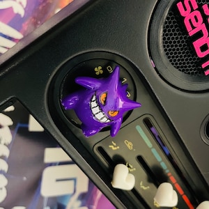 May include: A car dashboard with a purple cartoon character on a knob. The knob has numbers 0, 1, 2, and 3. The character has a wide smile and sharp teeth. The dashboard has a white heart-shaped knob and a black speaker with the word "sendai" printed on it.