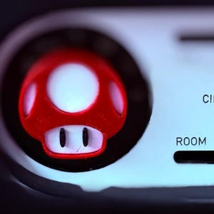 Może przedstawiać: Zbliżenie czerwono-białego guzika w kształcie grzyba, przypominającego postać z gry wideo, na jasnoszarym tle. Słowo „ROOM” jest widoczne pod guzikiem.