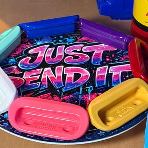 Op de afbeelding: Een ronde schaal met een zwarte achtergrond en blauw-roze graffiti-tekst "JUST SEND IT", omringd door diverse kleurrijke, rechthoekige kunststof handgrepen in de kleuren roze, geel, blauw, paars en wit.