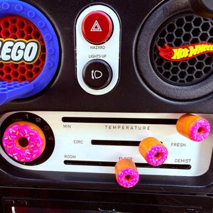 May include: Car dashboard with custom modifications. Features a blue LEGO-themed air vent, a red hazard light button, and a Hot Wheels logo. The temperature controls are adorned with donut-shaped knobs, adding a playful touch.