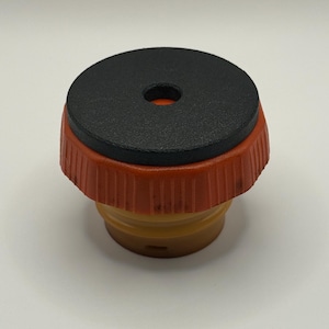 Peut inclure: Un bouchon de bouteille noir et orange avec un dessus rond noir et un petit trou au centre.
