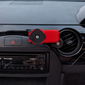 Può includere: Supporto per telefono da auto rosso e nero fissato al cruscotto di un'auto. Il supporto è posizionato vicino alla presa d'aria e presenta un supporto magnetico circolare. La parola "PUSH" è visibile.