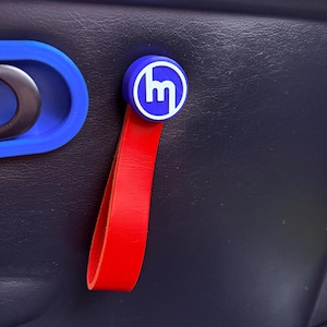 Puede incluir: Una tira de cuero roja para la manija de la puerta de un coche con un logotipo de Mazda azul y blanco.