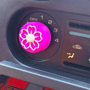 Puede incluir: Una perilla de coche rosa con un diseño de flor blanca. La perilla tiene los números del 1 al 4 y la palabra "OFF" impresa en ella.