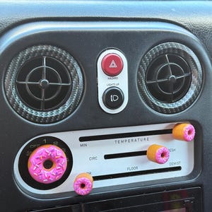 Puede incluir: Detalle interior de coche con salpicadero negro, rejillas de ventilación, botón de luces de emergencia y controles de temperatura. Perillas y detalles en forma de donut rosa añaden un toque lúdico al diseño interior del coche. El texto "HAZARD LIGHTS UP" y "TEMPERATURE" es visible.