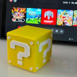 May include: A yellow cube featuring white question marks, inspired by a popular video game. The cube is positioned in front of a gaming console screen, which displays several game titles.