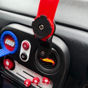 Puede incluir: Un soporte rojo para teléfono con base negra está fijado a un salpicadero negro. El salpicadero tiene detalles en rojo y blanco, incluyendo perillas en forma de hongo y un logotipo de Hot Wheels.