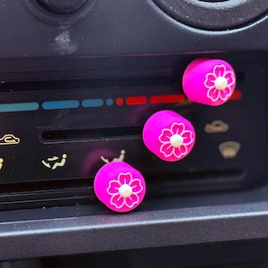 Może przedstawiać: Trzy różowe pokrętła z białymi wzorami kwiatów są przymocowane do czarnej deski rozdzielczej samochodu. Pokrętła służą do sterowania klimatyzacją samochodu.