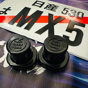 Könnte beinhalten: Zwei schwarze Kunststoff-Ventilkappen mit der Aufschrift "Turn Block Wise". Die Kappen liegen auf einer Oberfläche mit einem weißen Nummernschild mit schwarzer Schrift, auf dem "日産 530 MX5" steht.