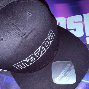 Puede incluir: Gorra de béisbol negra con un logotipo "Mazda" blanco en la parte delantera y una etiqueta "Snapback Original Headwear" blanca en el lateral.