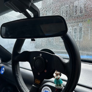 May include: A black leather-wrapped steering wheel with a black OMP logo and a small doll figurine in the centre. The steering wheel is in a car interior with a rearview mirror above it.