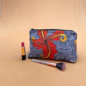 May include: A grey zipped makeup bag with a red and yellow floral design. The bag is sitting on a brown surface with a lipstick and a makeup brush.