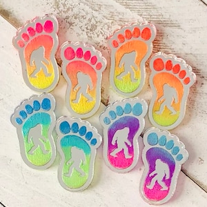 Könnte beinhalten: Acht bunte, acrylfarbene Fußabdruck-förmige Pins mit einer Silhouette von Bigfoot in jedem. Die Pins sind in einem Regenbogen-Farbverlauf, beginnend mit Rot und endend mit Lila.