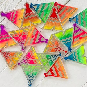 May include: A collection of 15 colorful, iridescent, and patterned teepee-shaped ornaments. The ornaments are made of a clear material with a holographic finish. The ornaments are arranged in a random pattern on a white wooden surface.