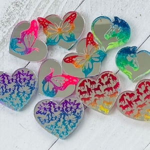 May include: A collection of heart-shaped mirrored acrylic charms. Some feature butterfly designs in vibrant colors, while others showcase horse silhouettes. The charms are arranged on a white wooden surface, creating a visually appealing display.