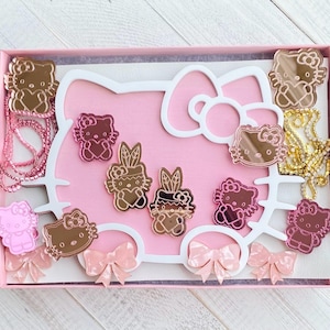 May include: A pink and white Hello Kitty themed jewelry box with a mirror and a variety of pink and gold Hello Kitty charms. The box is decorated with bows and hearts.