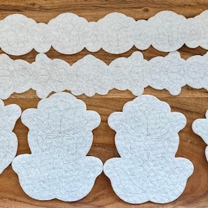 May include: A collection of white, die-cut felt shapes, including cloud-like borders, floral designs, and circular and stylized shapes. The pieces are arranged on a wooden surface, with white flowers in the upper right corner.