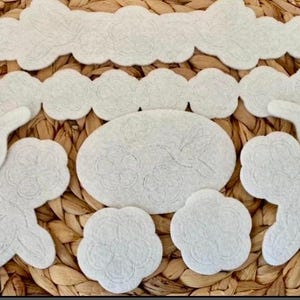 May include: A collection of light gray felt shapes, including cloud and floral designs, arranged on a woven wicker surface. The shapes vary in size and style, suitable for crafting or decorative purposes.