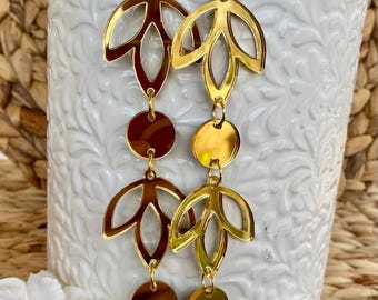 May include: Gold-colored dangle earrings with a leaf and circle design. The earrings are made of a shiny, reflective material and hang from gold-colored hooks. The earrings are displayed in front of a white vase with a floral pattern.