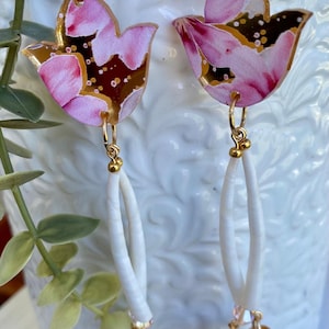 Könnte beinhalten: Goldfarbene Ohrringe mit floralem Design. Der obere Teil der Ohrringe hat ein rosa-braunes Blumenmuster. Sie haben einen weißen, oval geformten Anhänger. Die Ohrringe sind vor einem weißen Hintergrund abgebildet.