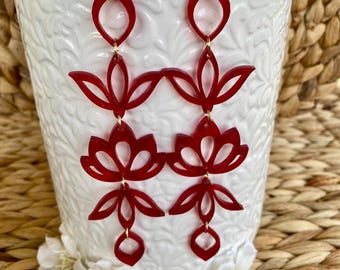 May include: A pair of long, dangling earrings featuring a deep red, translucent, laser-cut acrylic design. Each earring consists of three stacked, symmetrical floral-inspired geometric shapes connected by small gold-toned metal jump rings.