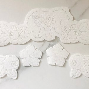 May include: A set of white felt cutouts featuring a bear, flowers, and berries. The cutouts are ideal for crafting projects such as cards, scrapbook pages, or home decor.
