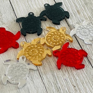 Könnte beinhalten: Sortiment von sieben schildkrötenförmigen Acryl-Anhängern in verschiedenen Farben, darunter Rot, Gold, Silber und Schwarz. Die Anhänger haben ein geometrisches Muster und sind perfekt, um Schmuck oder Bastelarbeiten eine Prise Verspieltheit zu verleihen.