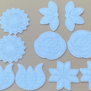 May include: Nine die-cut paper shapes in the form of flowers, including sunflowers, roses, and lilies. The shapes are white with a light blue tint and have a delicate, intricate design.