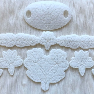 May include: A set of white felt flower appliques with embroidered details. The set includes a large flower with two blooms, a smaller flower with a single bloom, a long leaf garland, and a small oval shape with a single flower.