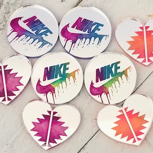 May include: Assortment of seven colorful, round and heart-shaped acrylic charms with a Nike logo and a geometric pattern. The charms are iridescent and have a rainbow-like effect.