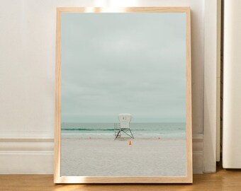 May include: A framed photograph of a lifeguard stand on a beach. The stand is white and has a number "14" on it. The beach is sandy and there are a few orange cones in the sand. The sky is cloudy and the ocean is blue.