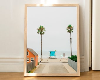 May include: A framed photograph of a beach scene with a blue lifeguard stand, two palm trees, and a small orange house in the distance. The beach is empty and the sky is a light blue.