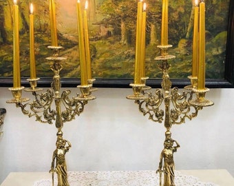 May include: A pair of gold metal candle holders with ornate designs and a female figure on each base. Each holder has six candle arms and is decorated with intricate scrollwork. The candle holders are displayed on a white lace tablecloth with a blue and white porcelain trinket box in the center.